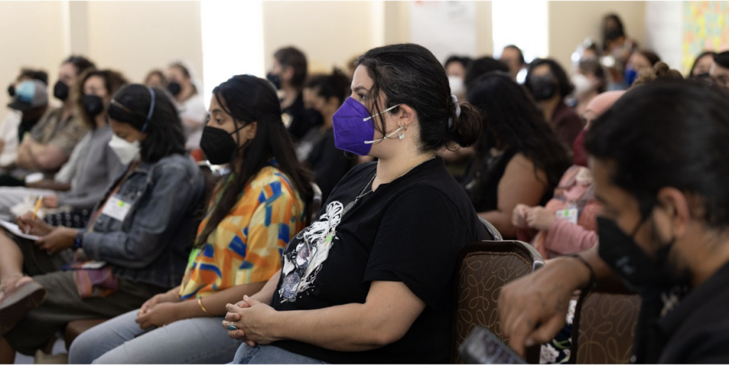 Feminine person with a light complexion and straight hair in a low ponytail wearing a deep purple N95 mask and black short sleeve shirt with white graphics. They are centered in the image but surrounded by people also wearing masks and seated very closely in a naturally lit auditorium.